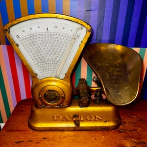 May include: An antique gold-colored Dayton scale with a white face and black numbers. The scale has a curved top with a dial and a brass-colored bowl. The base of the scale is rectangular and sits on a wooden surface.
