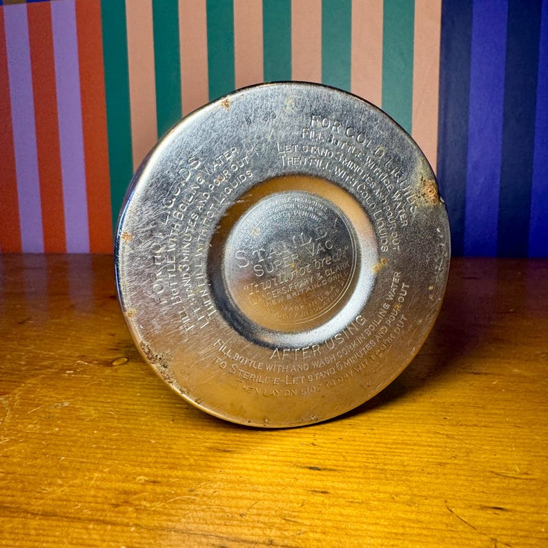 Pode incluir: Close-up da base de uma garrafa t&eacute;rmica Stanley Super Vac vintage. A base de metal mostra sinais de desgaste, com instru&ccedil;&otilde;es de uso em relevo. O texto inclui "Stanley Super Vac" e "It will not break".