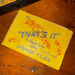 Op de afbeelding: Een vintage geel spiraalgebonden boek met de titel "THAT'S IT" met blauwe tekst en een rode drakenillustratie. Het boek, gedateerd 1943, gaat over het spelen van Chinese tegels. De omslag bevat ook illustraties van tegels.