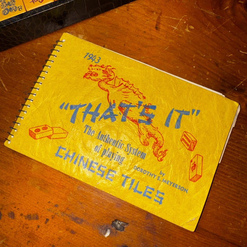 Op de afbeelding: Een vintage geel spiraalgebonden boek met de titel "THAT'S IT" met blauwe tekst en een rode drakenillustratie. Het boek, gedateerd 1943, gaat over het spelen van Chinese tegels. De omslag bevat ook illustraties van tegels.