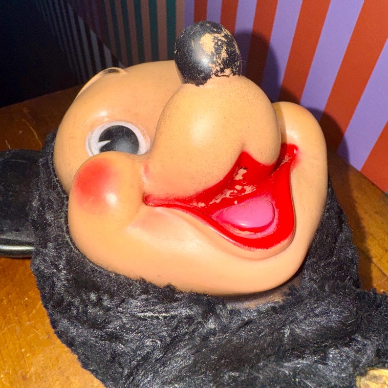 May include: Close-up of a vintage Mickey Mouse mask. The mask features a tan face with a black nose, black fur around the face, and a red mouth with a pink tongue. The mask is likely made of plastic or a similar material.