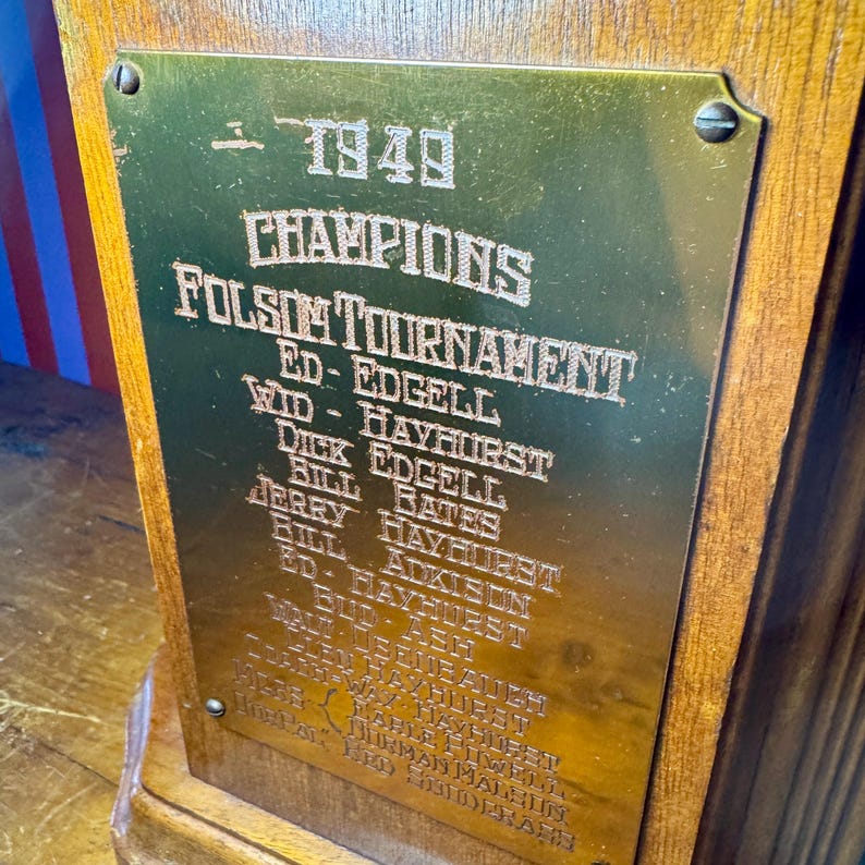 Puede incluir: Un trofeo de madera vintage con una placa dorada. La placa est&aacute; grabada con el texto "1949 CHAMPIONS FOLSOM TOURNAMENT" y enumera nombres. El trofeo est&aacute; montado sobre una base de madera.