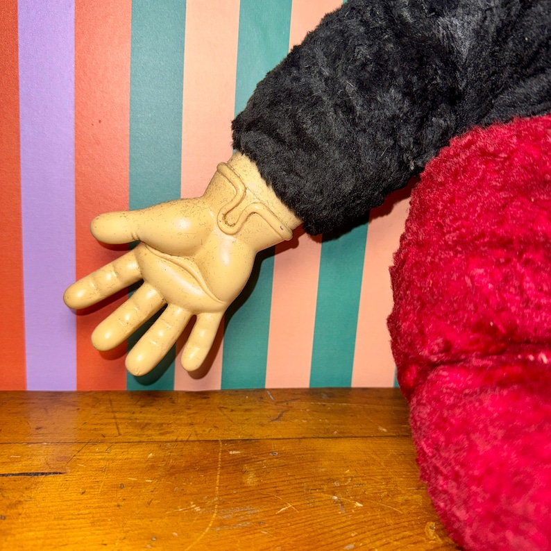 May include: A plush toy arm with a tan plastic hand, black fur sleeve, and red plush body. The hand is open, with fingers extended. The background features vertical stripes in orange, green, purple, and white. The toy rests on a wooden surface.