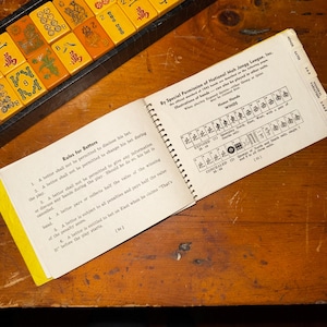 Op de afbeelding: Een open Mahjong-regelboek ligt op een houten tafel naast een bakje met tegels. Het boek toont tekst en diagrammen die handen en gokregels illustreren. De tegels zijn geel en oranje.