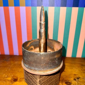 May include: A repurposed bullet stands upright in a copper-colored metal container. The container sits on a wooden base, with a colorful striped background. The bullet is a dark bronze color.