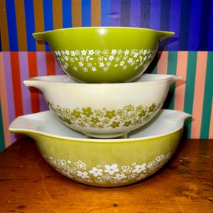 May include: Three vintage Pyrex mixing bowls in shades of green and cream, stacked. Each bowl features a white floral pattern around the rim. The bowls have a classic design with a pouring spout on each side.