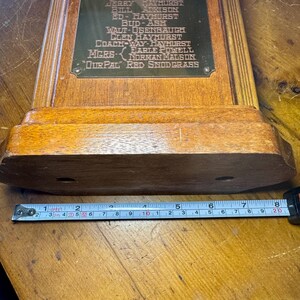 Puede incluir: Una base de trofeo de madera vintage con una placa de lat&oacute;n. La placa presenta nombres y texto grabados. Una cinta m&eacute;trica muestra que la base mide aproximadamente 20 cm de largo.