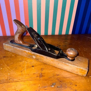 May include: A vintage wooden hand plane with a dark brown handle and knob. The plane has a black metal blade and is set on a rectangular wooden base. The tool is designed for woodworking and smoothing surfaces.