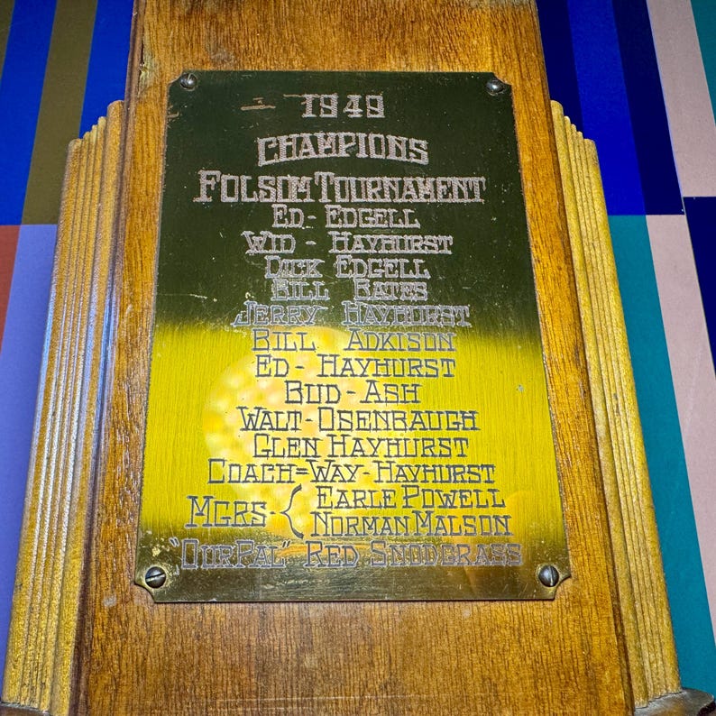 Puede incluir: Un trofeo de madera vintage con una placa dorada. La placa presenta el texto "1949 CHAMPIONS FOLSOM TOURNAMENT" junto con una lista de nombres. El trofeo tiene un acabado de madera oscura y est&aacute; montado sobre una base de madera.