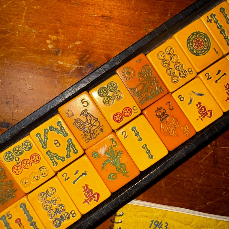 Op de afbeelding: Een vintage Mahjong-set met gele tegels met verschillende symbolen en karakters. De tegels zijn gerangschikt in een zwarte lade op een houten oppervlak. De set bevat tegels met nummers, pakken en illustraties.