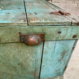 May include: A weathered wooden chest painted in shades of teal and green, with visible wood grain and distressed paint. Features a metal handle and hinges, showcasing an antique aesthetic. The chest's design suggests it could be used for storage.