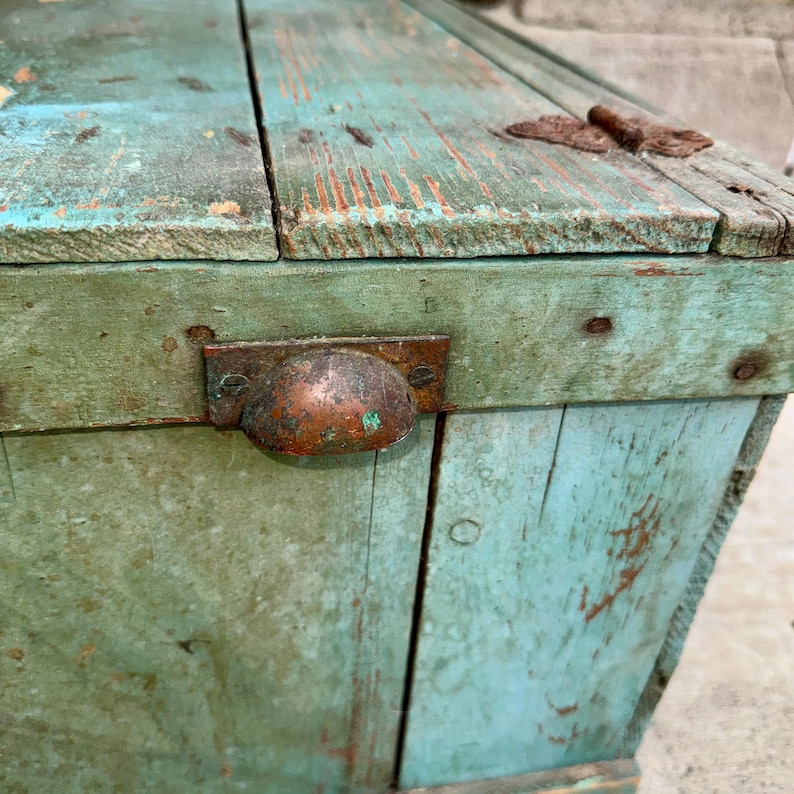 May include: A weathered wooden chest painted in shades of teal and green, with visible wood grain and distressed paint. Features a metal handle and hinges, showcasing an antique aesthetic. The chest's design suggests it could be used for storage.