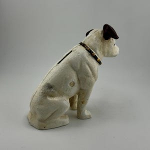 Op de afbeelding: Een vintage witte gietijzeren spaarpot in de vorm van een hond. De hond heeft bruine oren en een zwarte halsband met goudkleurige studs. De spaarpot heeft een gleuf aan de bovenkant voor het plaatsen van munten en een muntafgifte aan de onderkant. De hond zit.