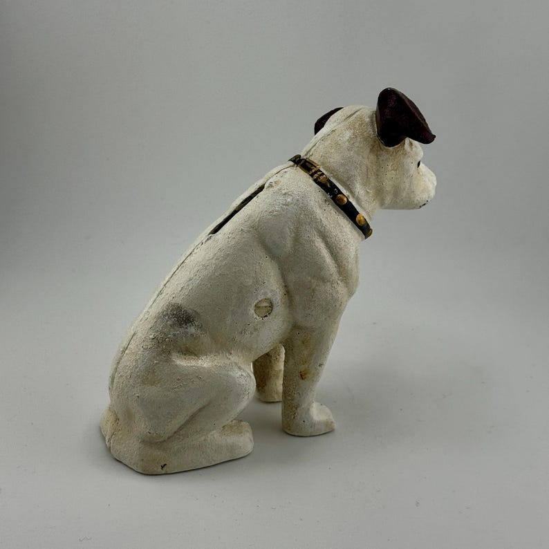 Op de afbeelding: Een vintage witte gietijzeren spaarpot in de vorm van een hond. De hond heeft bruine oren en een zwarte halsband met goudkleurige studs. De spaarpot heeft een gleuf aan de bovenkant voor het plaatsen van munten en een muntafgifte aan de onderkant. De hond zit.