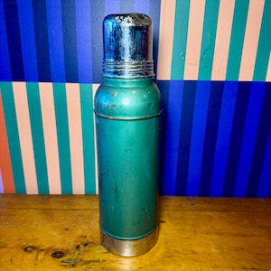 Pode incluir: Uma garrafa t&eacute;rmica de metal verde vintage com tampa e base prateadas. A garrafa t&eacute;rmica tem formato cil&iacute;ndrico e apresenta sinais de desgaste, sugerindo que &eacute; um item antigo. O fundo apresenta um padr&atilde;o listrado colorido.