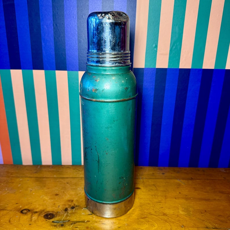 Pode incluir: Uma garrafa t&eacute;rmica de metal verde vintage com tampa e base prateadas. A garrafa t&eacute;rmica tem formato cil&iacute;ndrico e apresenta sinais de desgaste, sugerindo que &eacute; um item antigo. O fundo apresenta um padr&atilde;o listrado colorido.