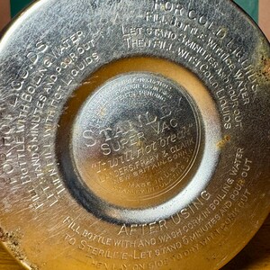 Pode incluir: Close-up da tampa de uma garrafa t&eacute;rmica Stanley Super Vac vintage. A tampa &eacute; prateada com texto em relevo detalhando as instru&ccedil;&otilde;es de uso para l&iacute;quidos quentes e frios. O texto inclui "It will not break" e "Made in U.S.A."