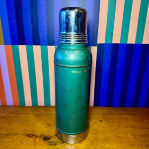 Pode incluir: Uma garrafa t&eacute;rmica verde vintage com tampa e base prateadas. A garrafa t&eacute;rmica &eacute; cil&iacute;ndrica e mostra sinais de desgaste. O fundo apresenta um padr&atilde;o de listras coloridas.