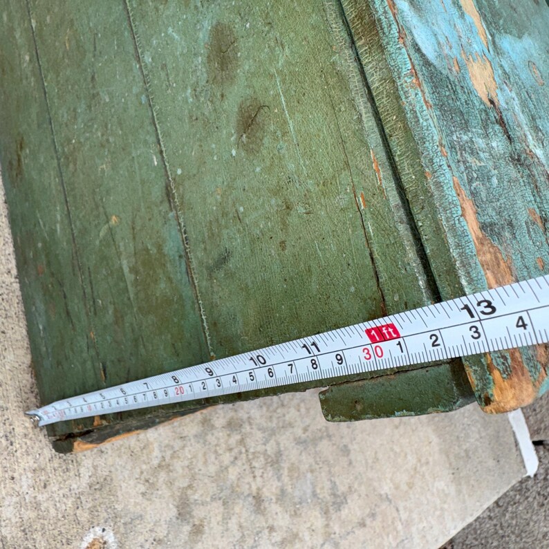 May include: Close-up of a distressed, vintage wooden box with peeling green and blue paint. The box is being measured with a tape measure, showing a length of approximately 30 inches.