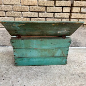 May include: A weathered, teal-painted wooden box with a hinged lid. The box is constructed from horizontal planks and shows signs of age and wear, with visible nail heads and distressed paint. The lid is angled, and the box sits on a concrete surface.