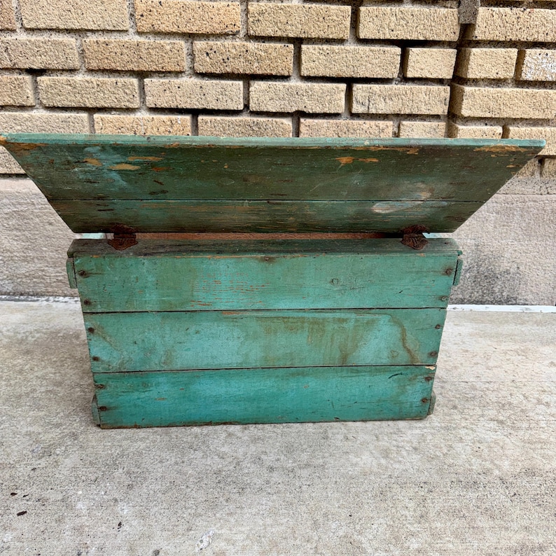 May include: A weathered, teal-painted wooden box with a hinged lid. The box is constructed from horizontal planks and shows signs of age and wear, with visible nail heads and distressed paint. The lid is angled, and the box sits on a concrete surface.