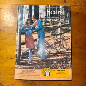 May include: Vintage Sears Fall/Winter 1976 catalog cover featuring a couple in matching blue sweaters and jeans, leaning on a wooden fence. Two bicycles are in the background. The Sears logo is at the top.