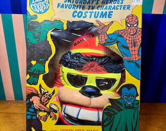 Puede incluir: Una caja de disfraz vintage Ben Cooper Saturday's Heroes Favorite TV Character con fondo amarillo e ilustraciones de personajes de cómic. Dentro de la caja hay una máscara de plástico de un personaje con pelo negro y gafas amarillas.