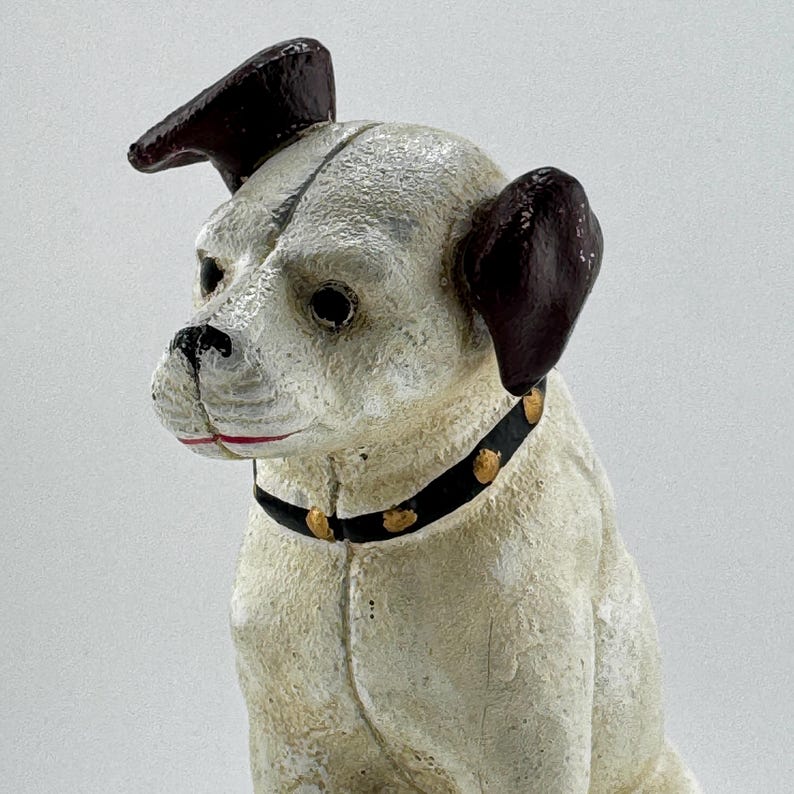 Op de afbeelding: Een vintage hondenbeeldje met een cr&egrave;mekleurig lichaam, donkerbruine oren en een zwarte halsband versierd met goudkleurige studs. De hond heeft een gedetailleerd, verouderd gezicht met een zwarte neus en rode lippen. Decoratief item.