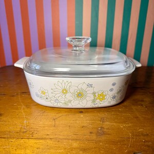 May include: A vintage casserole dish with a clear glass lid and a clear glass handle. The dish is white with a floral pattern in yellow, green, and blue. The dish is on a wooden surface.