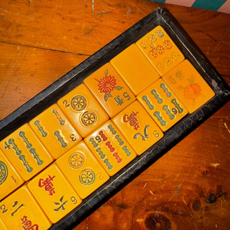 Op de afbeelding: Een vintage Mahjong-set met gele tegels en groene en rode markeringen, gerangschikt in een zwarte lade. De tegels bevatten verschillende symbolen en cijfers, waaronder bloemen en Chinese karakters. De set wordt gepresenteerd op een houten oppervlak.