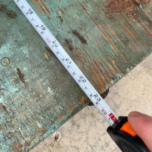 May include: A close-up of a worn, teal-colored wooden surface with a metal tape measure extended across it. The tape measure shows measurements in inches and feet. The wood has visible wear and tear.