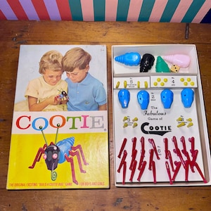 May include: Vintage Cootie board game with a colorful box featuring children and a cartoon bug. The game includes plastic bug parts and dice. The box reads "The Fabulous Game of Cootie" and "Build a Cootie Bug".
