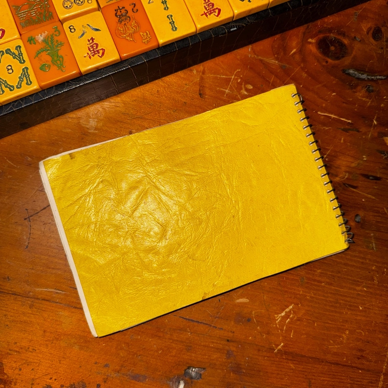 Op de afbeelding: Een geel spiraalgebonden notitieboek met een getextureerde kaft ligt op een houten oppervlak. De pagina's van het notitieboek zijn wit en de spiraalbinding is zilverkleurig. Op de achtergrond is een deel van een Mahjong-set te zien.