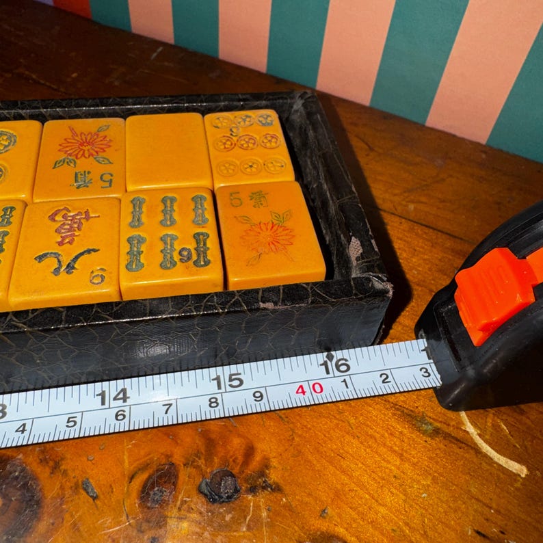 Op de afbeelding: Een vintage Mahjong-set met oranje tegels met Chinese karakters en bloemmotieven, weergegeven in een zwarte koffer. De set wordt gemeten met een meetlint, ongeveer 40 cm lang.