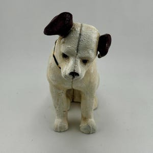 Op de afbeelding: Een vintage hondenbeeldje in zittende positie. De hond heeft een cr&egrave;mekleurig lichaam, donkerbruine oren en een zwarte halsband. Het beeldje heeft een verweerde, getextureerde uitstraling, wat suggereert dat het een antiek of een replica is.