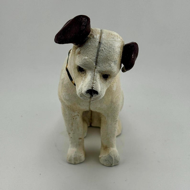 Op de afbeelding: Een vintage hondenbeeldje in zittende positie. De hond heeft een cr&egrave;mekleurig lichaam, donkerbruine oren en een zwarte halsband. Het beeldje heeft een verweerde, getextureerde uitstraling, wat suggereert dat het een antiek of een replica is.