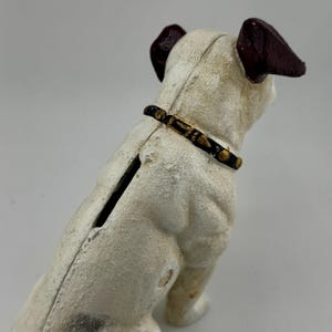 Op de afbeelding: Een vintage witte gietijzeren spaarpot in de vorm van een hond met bruine oren en een zwarte halsband met gouden accenten. De hond zit rechtop, met een gleuf op zijn rug om munten in te doen. De spaarpot heeft een getextureerd, verouderd uiterlijk.