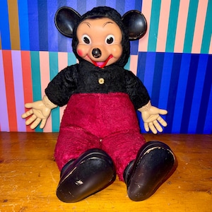 May include: Vintage Mickey Mouse plush doll. The doll features a black hooded jacket with a gold button, red pants, and large black shoes. The doll has a plastic face with large eyes, a red smile, and black ears. The doll is sitting on a wooden surface.