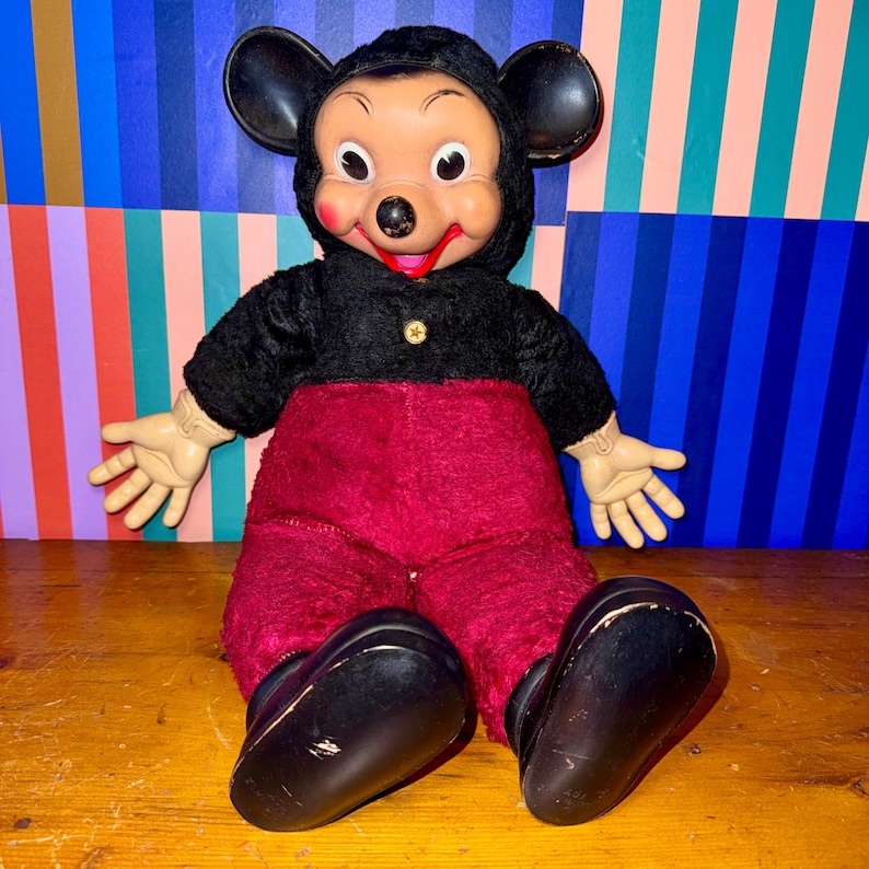 May include: Vintage Mickey Mouse plush doll. The doll features a black hooded jacket with a gold button, red pants, and large black shoes. The doll has a plastic face with large eyes, a red smile, and black ears. The doll is sitting on a wooden surface.