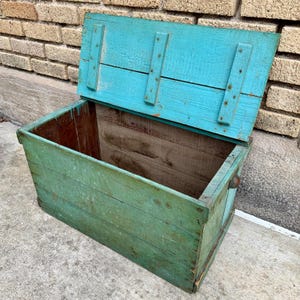 May include: An open, weathered wooden chest painted in shades of teal and green. The lid is propped open, revealing the interior. The chest has metal hardware and is made of horizontal wooden planks.