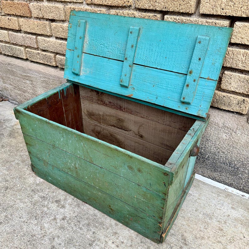May include: An open, weathered wooden chest painted in shades of teal and green. The lid is propped open, revealing the interior. The chest has metal hardware and is made of horizontal wooden planks.