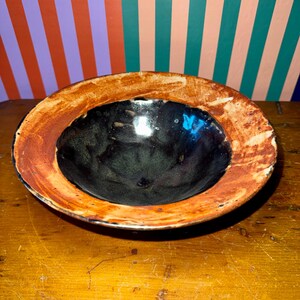 May include: A handmade ceramic bowl with a dark interior and a wide, textured rim. The bowl's interior is a glossy black, contrasting with the reddish-brown and cream-colored rim. The bowl is set on a wooden surface.
