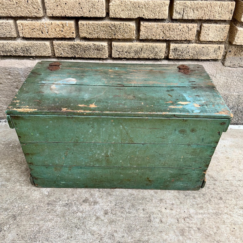 May include: A weathered, green wooden storage chest with a hinged lid. The chest has visible wood grain and signs of wear, including chipped paint and rust on the hinges. The rectangular box is set against a brick wall and concrete surface.