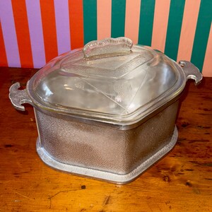 May include: A clear glass casserole dish with a lid, set on a wooden surface. The dish has a textured, silver-colored metal base and handles. The lid features a decorative handle and etched floral designs.
