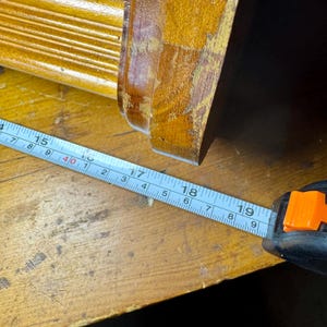 Puede incluir: Primer plano de un mueble de madera con una cinta m&eacute;trica que muestra una medida de aproximadamente 45,7 cm. El mueble es de color marr&oacute;n dorado c&aacute;lido, y la cinta m&eacute;trica es azul y blanca.