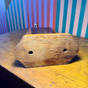 Puede incluir: Un antiguo molinillo de caf&eacute; de madera con cuerpo rectangular y parte superior decorativa. La base es de color marr&oacute;n claro con dos agujeros circulares. El molinillo tiene un mango de lat&oacute;n y est&aacute; sobre una superficie de madera.