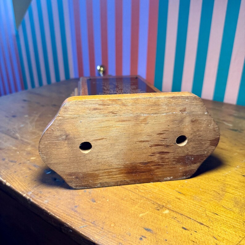 Puede incluir: Un antiguo molinillo de caf&eacute; de madera con cuerpo rectangular y parte superior decorativa. La base es de color marr&oacute;n claro con dos agujeros circulares. El molinillo tiene un mango de lat&oacute;n y est&aacute; sobre una superficie de madera.