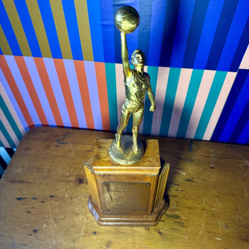 Puede incluir: Un trofeo de baloncesto dorado vintage que representa a un jugador sosteniendo un bal&oacute;n de baloncesto por encima de su cabeza. El trofeo est&aacute; montado sobre una base de madera con una placa. El fondo tiene un patr&oacute;n geom&eacute;trico colorido.
