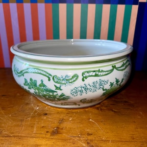 Könnte beinhalten: Eine weiße Keramikschale mit einem grün-weißen Dekor. Die Schale hat einen breiten Rand und eine abgerundete Form. Das grüne Muster zeigt Bäume, Vögel und andere natürliche Elemente. Die Schale steht auf einer Holzoberfläche.