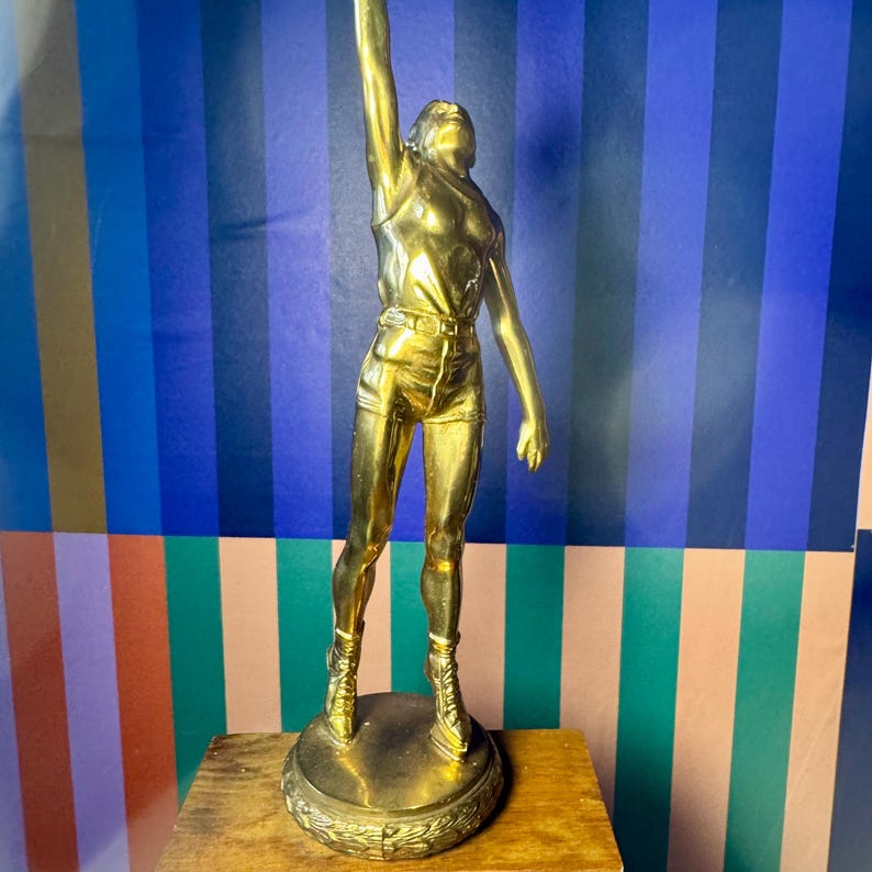 Puede incluir: Una estatua dorada de una persona con ropa deportiva, con un brazo levantado. La figura lleva pantalones cortos y una camiseta sin mangas. La estatua est&aacute; montada sobre una base circular con un borde decorativo. El fondo presenta un patr&oacute;n de rayas de colores.